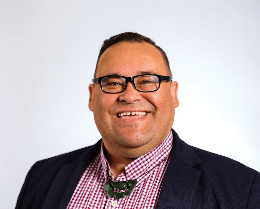 a man with light brown skin, a gingham red shirt, glasses, a big ponaumu and a smile in front of a plain background