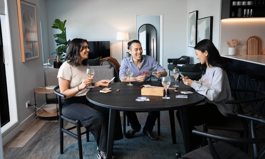 Three friends are sitting around a table in a cozy living room, playing a card game and enjoying drinks. The room has modern decor with framed artwork on the walls and a large plant in the corner.