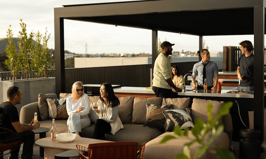 A group of six people socializing on a rooftop terrace. Three are sitting on a couch with drinks, while the other three are standing and talking near an outdoor kitchen area. The background shows a scenic view of a cityscape.