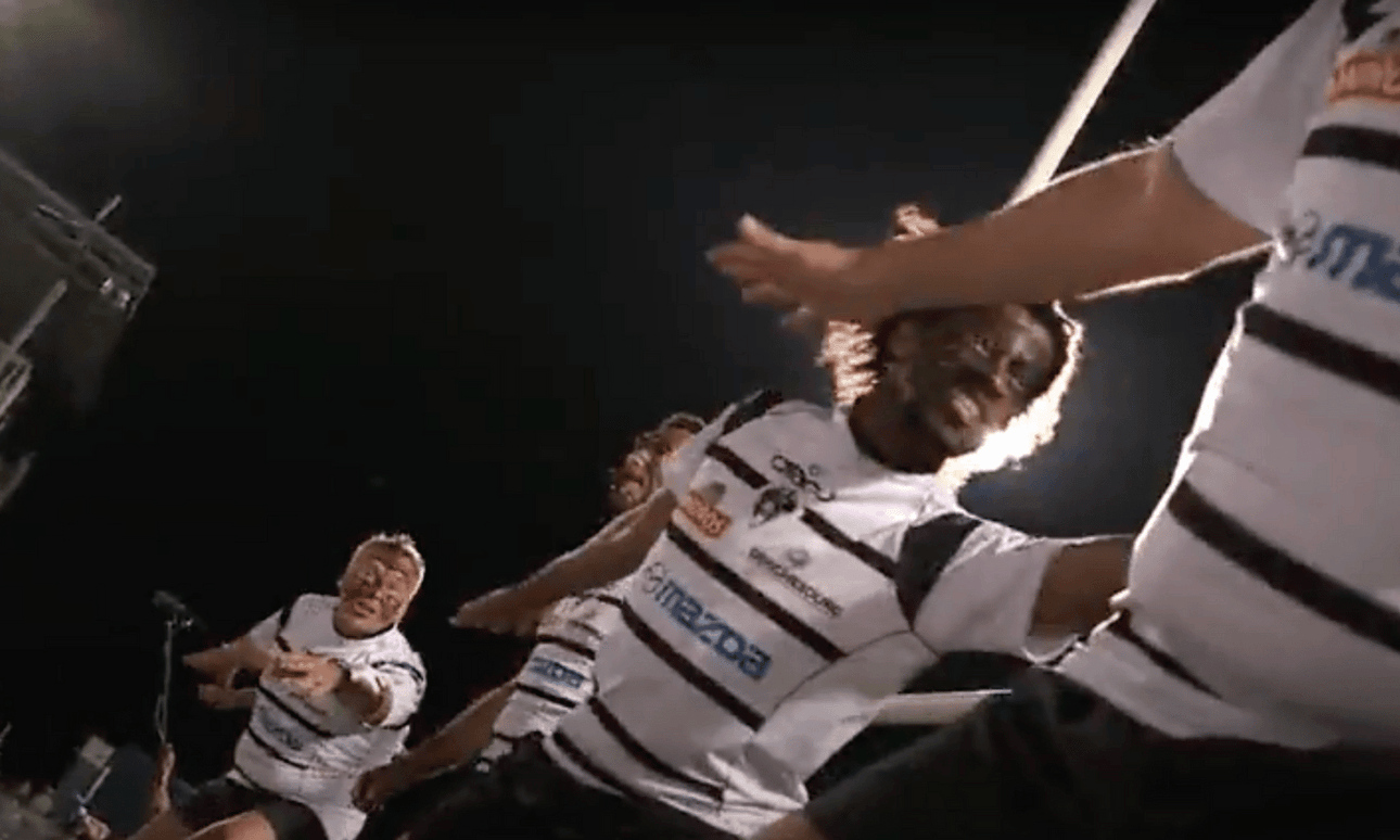 A group of four men dressed in white rugby uniforms with imitation mataora lead a haka.