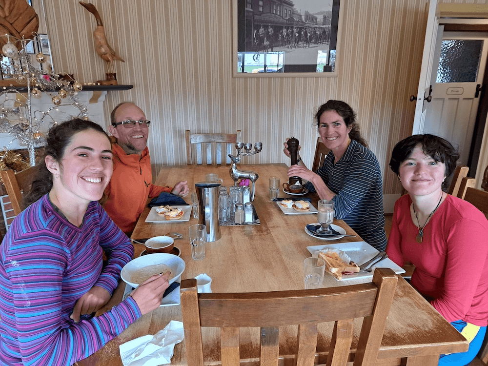 a family sitting at a table, two adults and two kids, all with big families. Mum emily is wearing a ratty stripey polypro shirt at the back right, and daugther ngaire is wearing, blue, pink and purple stripes at the front left