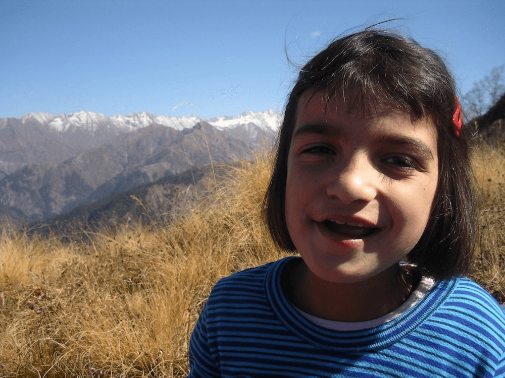 a brown-skinned, gormless looking small child with bright red plastic clips in her short hair, looks at the camera. Big himalayan mountains are in the background, and she is wearing a bright blue striped synthetic long sleeve top