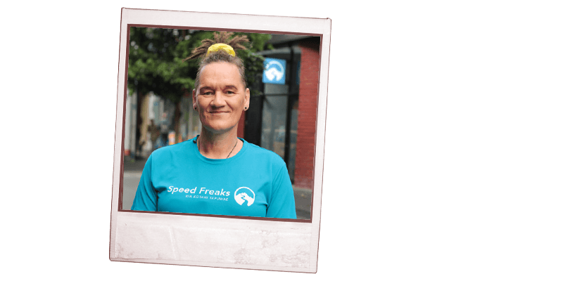 Simone Winter with a yellow scrunchie in their hair and wearing a blue 'Speed Freaks' t-shirt is smiling and standing on a city street.