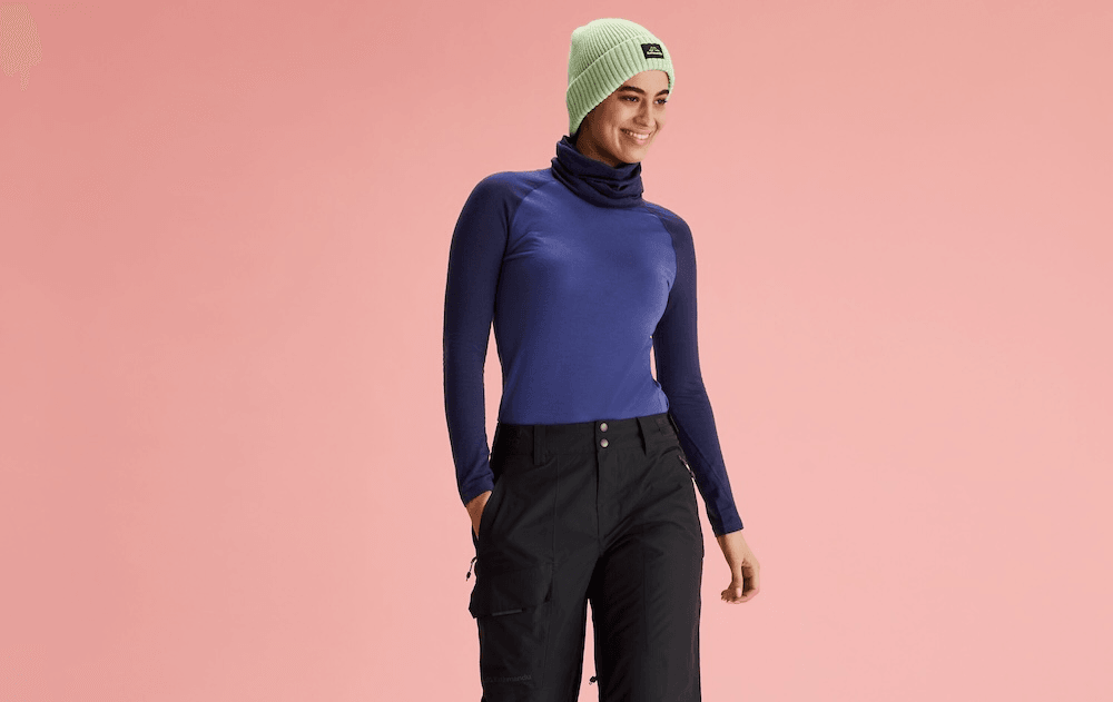 a plain pink background and famile model wearing black pants, a dark blue shirt and a light green Kathmandu beanie