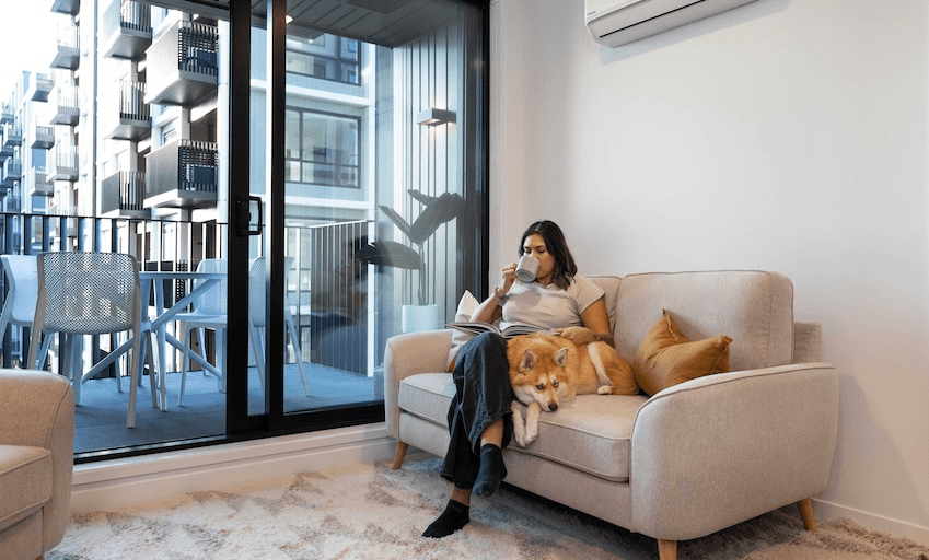 A woman is sitting on a sofa in a bright living room, sipping a drink and reading a book. Next to her is a dog resting comfortably on the couch. A sliding glass door opens to a balcony with outdoor furniture, showing apartment buildings outside.