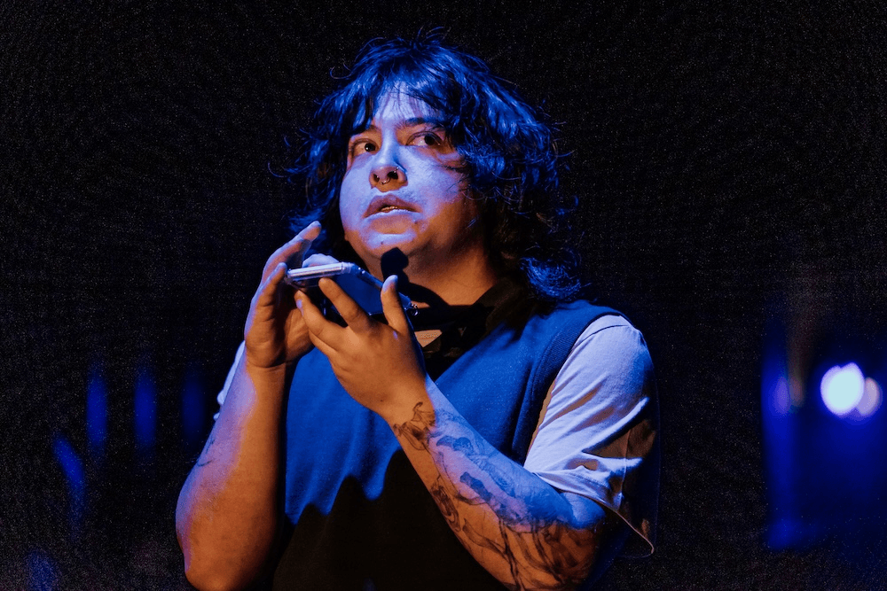 a Filopino/pakehā man with a full-arm tattoo sleeve stands in blue-toned lighteing, holding a phone and looking anxious