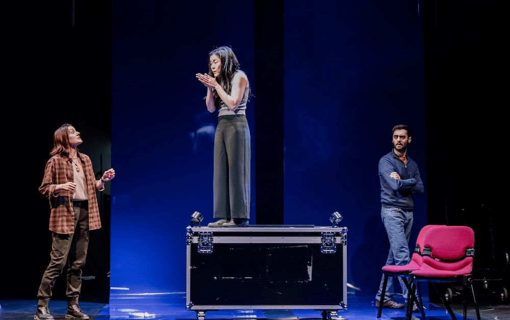 Dawn Cheong, a short Asian woman wearing flowy grey pants and a sleeveless top stands on a black box while two characters on each side of the blue-lit stage look on