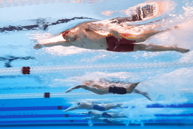 a clear blue pool with an underwater view and athletes on the surve with their limbs outstretched and ripples echoeing out from their bodies.