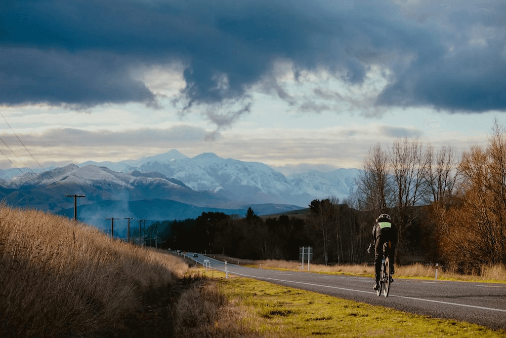 big south island mountains and a long gray road, with wilcox on a bike in a neon vest in the centre of the frame 