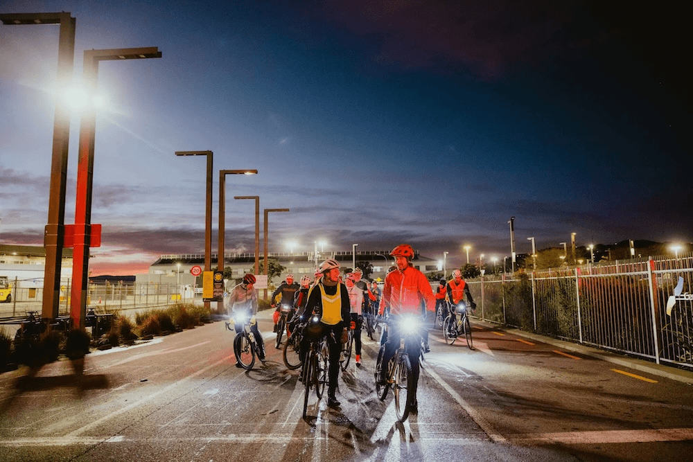 a dark morning with wellington harbour in the background and a big group of people with lights on their bikes travelling along a road