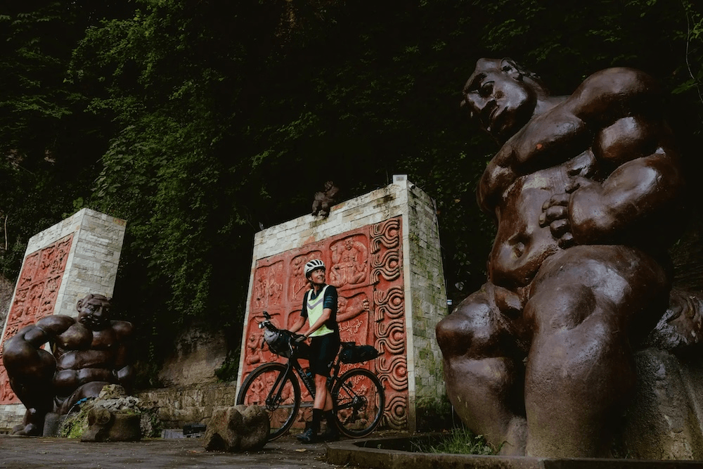 a woman with a bike and packs strapped to it, with a red tiled textured sculpture in the backround