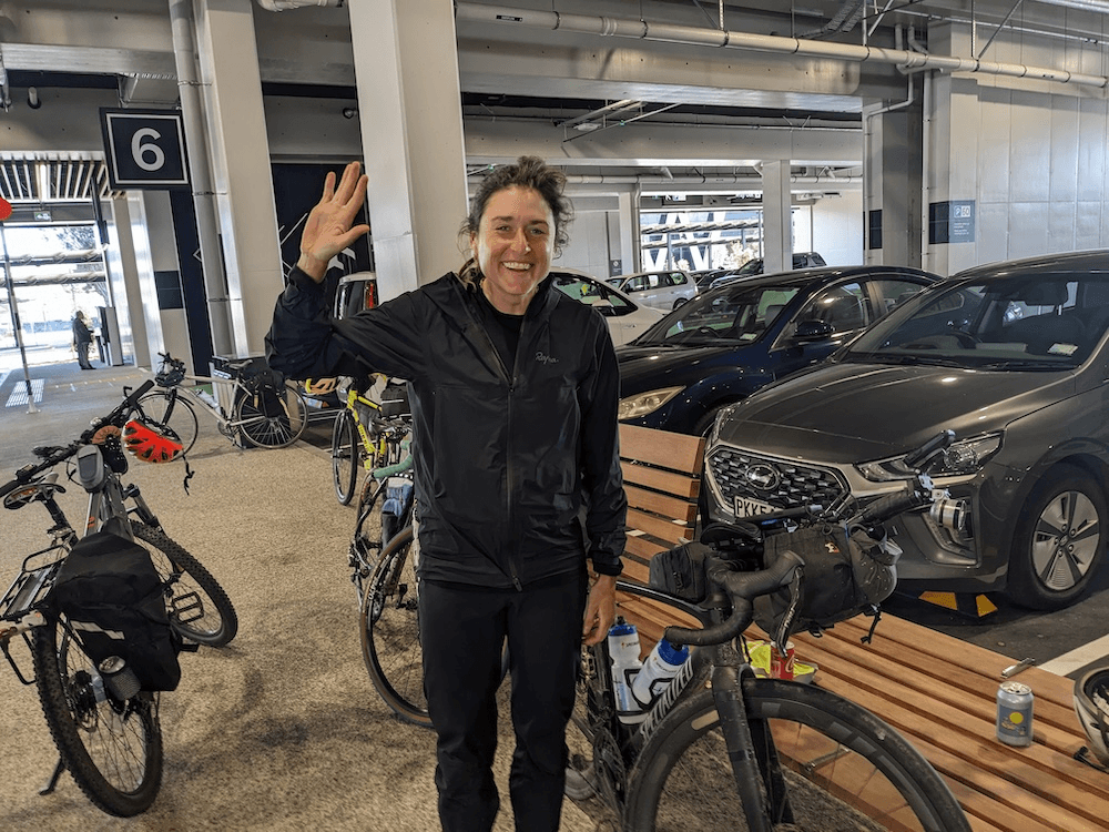 Lael wilcox, a woman in her thirties whit her hair in a scappty ponytail, wearing black outdoor gear, smiles while standing next to her bike, with an airport carpark in the background