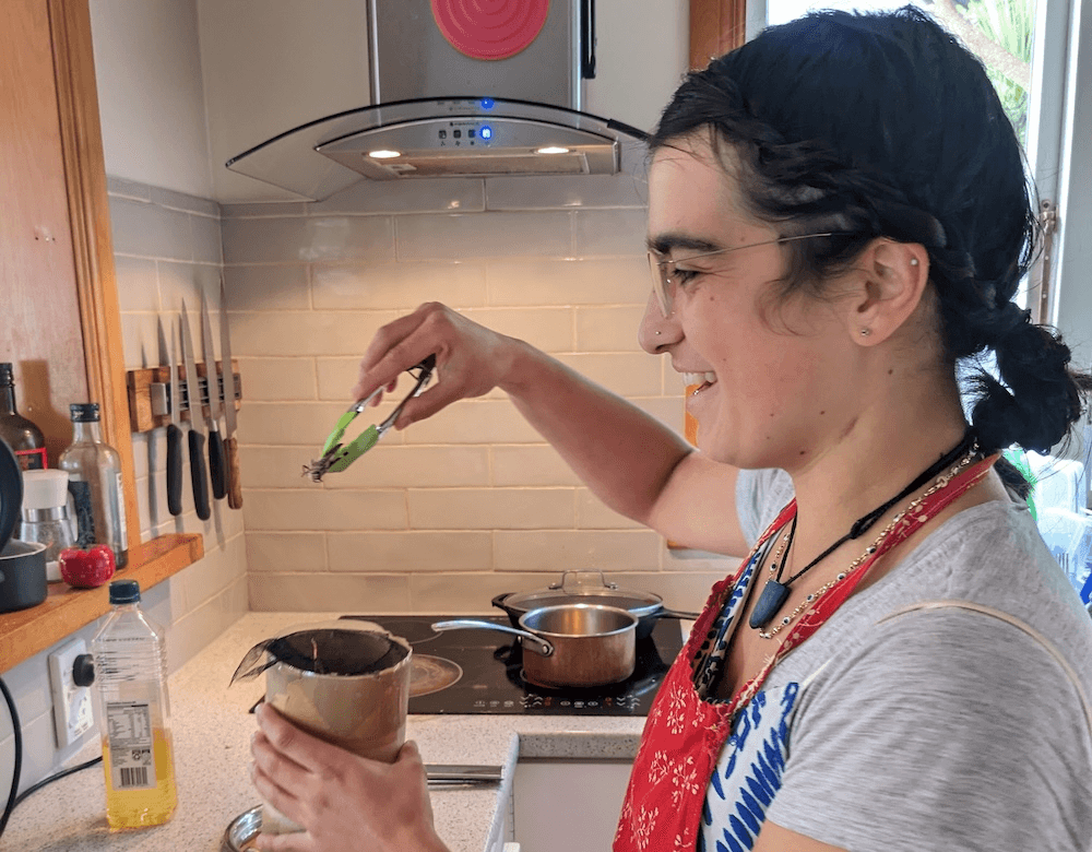 Shanti, a woman with light brown skin, glasses, and a grey t-shirt, grins nervously as she pulls a frozen locust out of a cardboard tube with a pair of green tongs. The stove with a hot, oily pan waiting in the background is a sign of the locust's eventual fate 