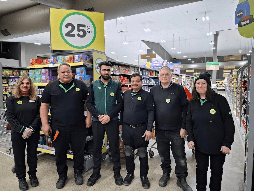 workers in black and green woolworths uniforms with a supermarket in the background, with conspicuous stickers on top of their uniforms