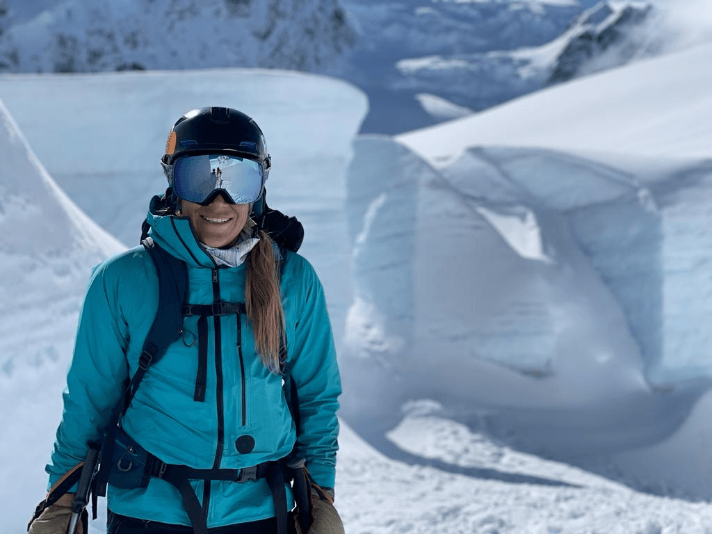 a white woman with blonde hair you can only see the lower part of her face because there is so much snow in the background and she is wearing reflective goggles and a light blue jacket