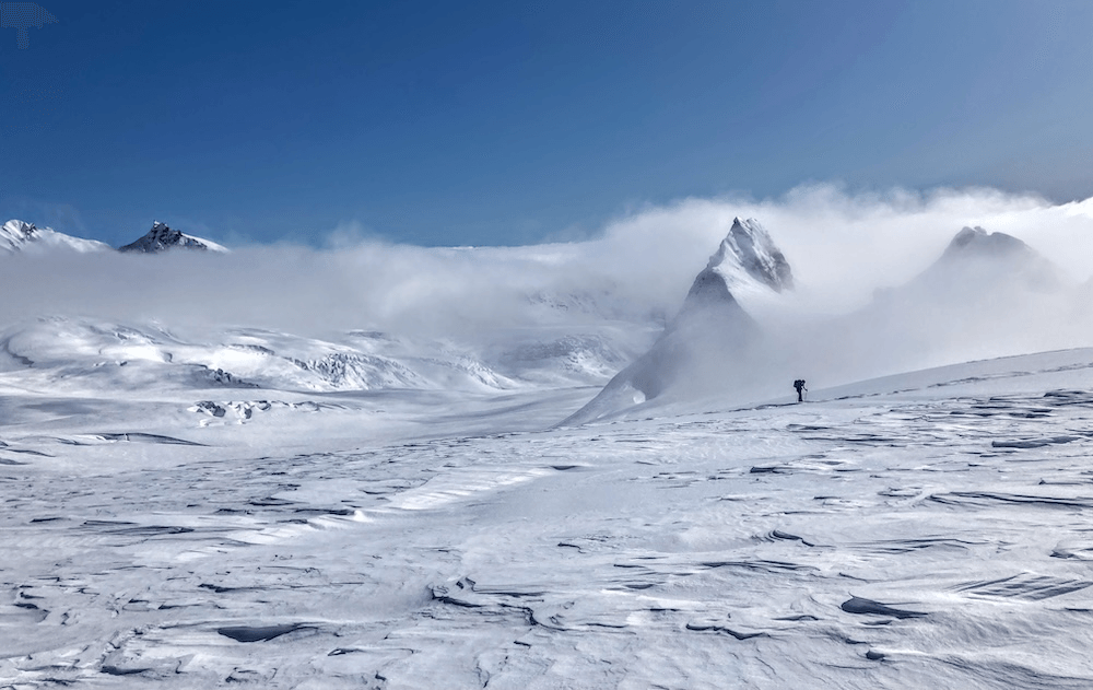 a snowy day in the mountains that makes you wish you had crampons on and were in the big wide world not looking at a screen; the sky is clear and blue, little cloaks of cloud are draped around the maunga, and a figure walks through a snow field in the sunshine