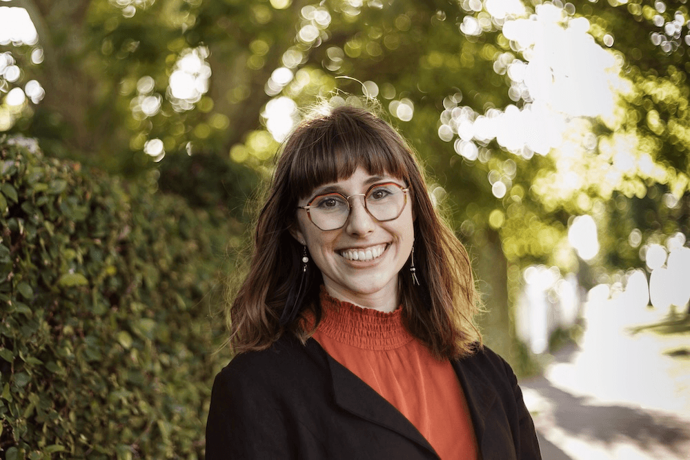 a woman with brown hair and pale skin wearing a huge smile and big glasses, a dork orange shirt and black jacket with trees in the background