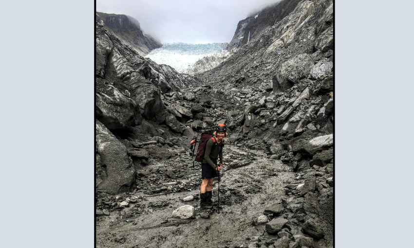 a white many with a silly expression on his face stands on a very muddy, rocky bit of land with a retreating tongue of glacier in background