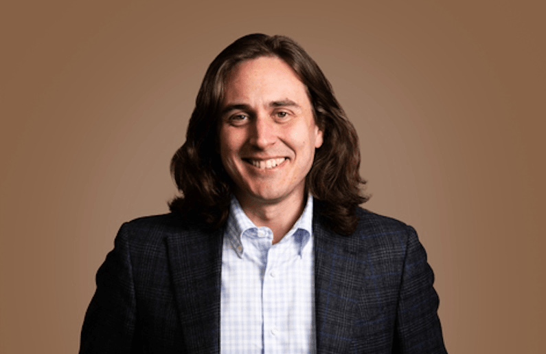 a brwon background with a white man with glossy long dark brown hair wearing a suit jacket and shirt and smiling at the camera