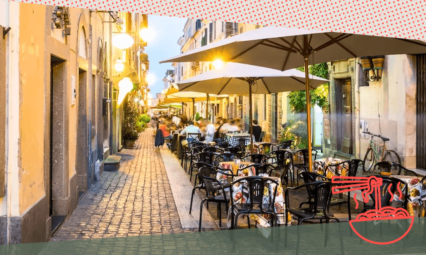 A cozy outdoor café in a European street with cobblestones and warm lights. Empty chairs and tables with colorful cushions line the walkway. People are dining under large umbrellas against a backdrop of charming, rustic buildings.