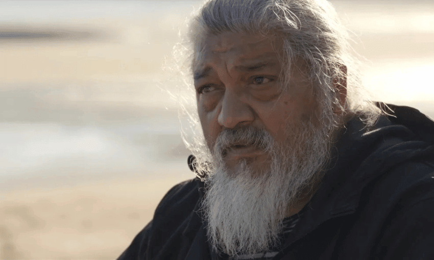 A man with long, gray hair and a beard is looking towards the camera. The background is a blurred outdoor setting with muted colours, indicating a beach or open area. The lighting suggests it might be late afternoon.