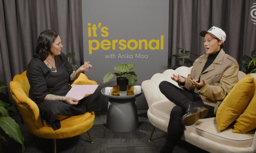 Two people, Anika Moa and Kiri Allan, sit on yellow and white chairs in a cozy setting, engaged in a conversation. A sign reads "it's personal with Anika Moa" against a gray backdrop. The scene is casual with plants and small tables around them.