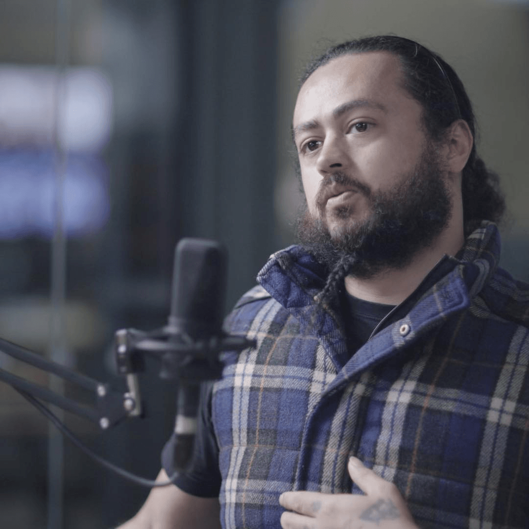 A man with a beard and long hair speaking into a microphone, wearing a blue flannel shirt.