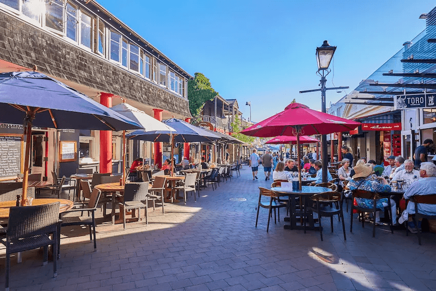 A lively outdoor cafe scene in Queenstown with people dining under colourful umbrellas on a sunny day. The street is lined with shops and restaurants, and a clear blue sky can be seen overhead.