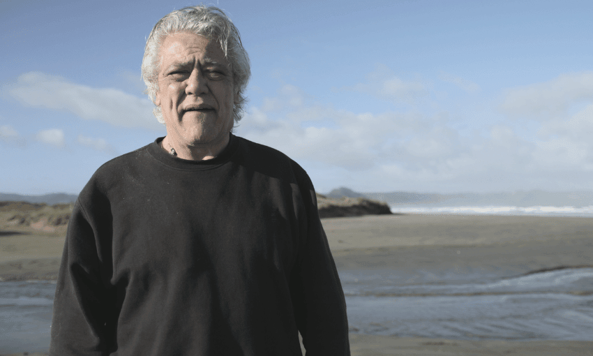 Haami Piripi standing on the beach in front of a creek with a mountain and the blue sky in the background.