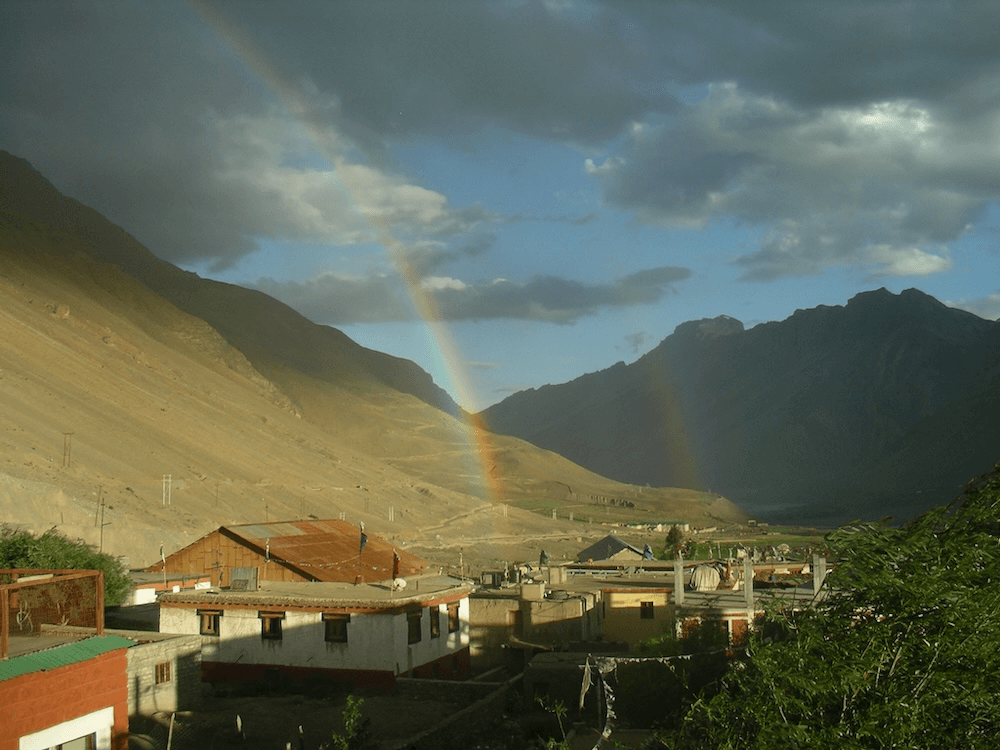 a valley with big mountains and mud brick houses with rainbows arching over them