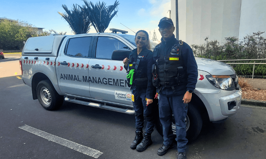 Two people in uniforms stand in front of a silver truck labeled "Animal Management." They are smiling and holding leashes. The setting appears to be an outdoor urban area with trees and a building.