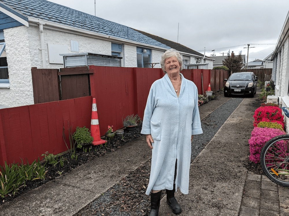 Faye, a 79 year old woman in a pastel blue dressing gown, stands on her driveway wearing gumboots