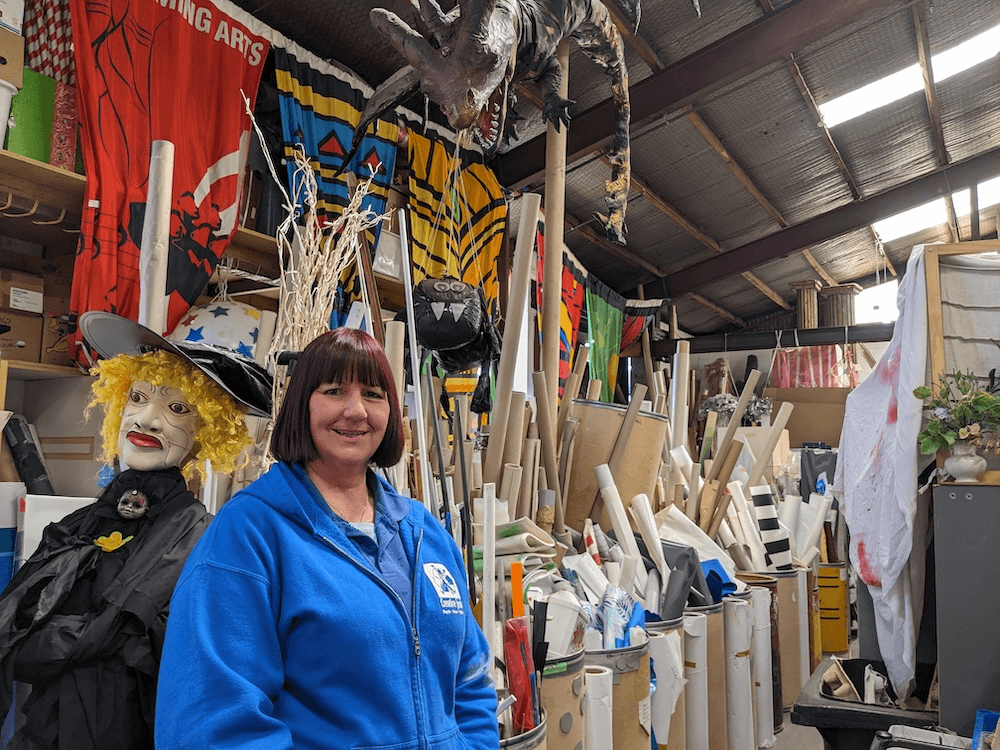 Christine Gayton, a middle aged woman with dyed red hair and a bright blue jacket smiles at the camera with a delightful chaos of cardboard tubes, a mannequin witch, and a giant dinosaur behind her