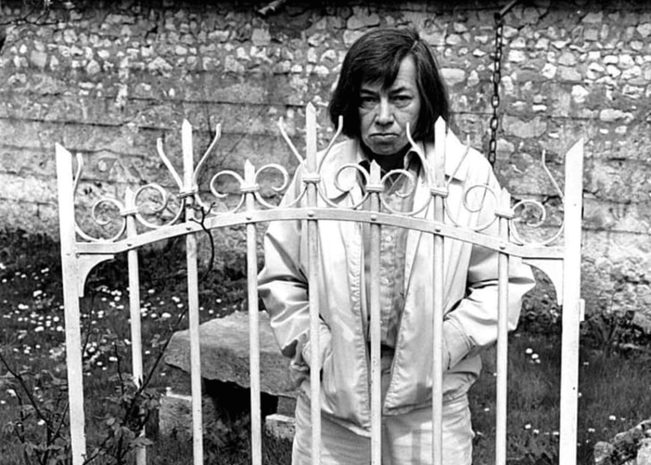 Patricia Highsmith stands behind a wrought iron gate, glaring in the camera, her hands in the pockets of her jacket.