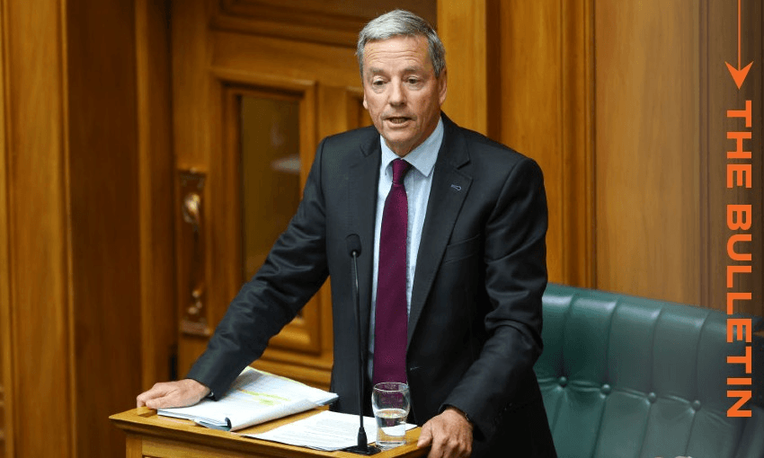 A man in a suit stands at a lectern with papers and a glass of water in front of him. He appears to be speaking in a wooden panelled room. The text "The Bulletin" is vertically displayed on the right side of the image.