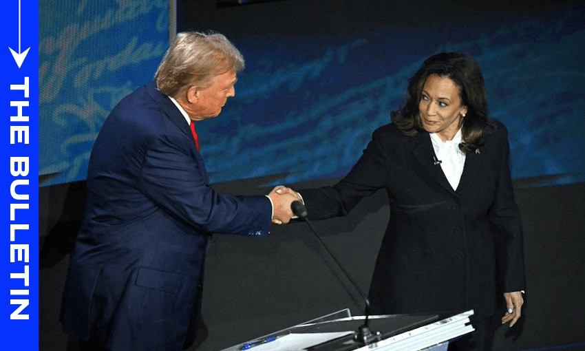 Two people in formal attire shake hands on stage during a debate. A digital banner on the left reads "THE BULLETIN." The setting suggests a public or political event with podiums and stage lighting.