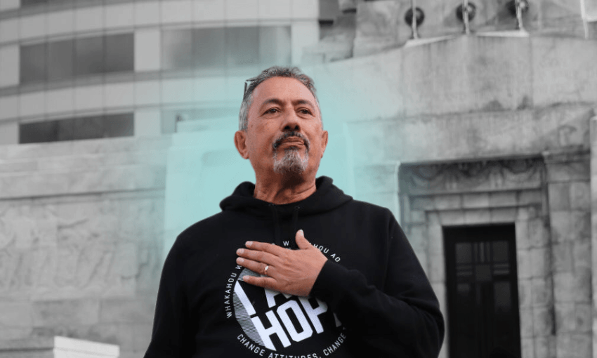 Mike King wears a black hoodie and stands in front of parliament buildings in wellington with his hand on his chest