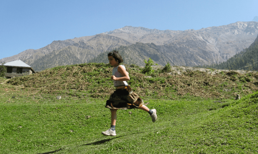 a small brown girl wearing a brown skirt and running shoes, is slightly out of focus as she strides across a grassy path, with big mountains and a blue sky in the background