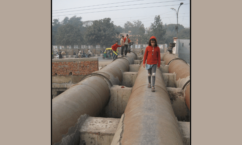 A SMALL GIRL WITH PATCHES ON HER STRIPED LEGGINS WEARING A SOFT ORANGE HOODED SWEATER WALKS AcROSS a pipe with polluted air and some men standing in the background