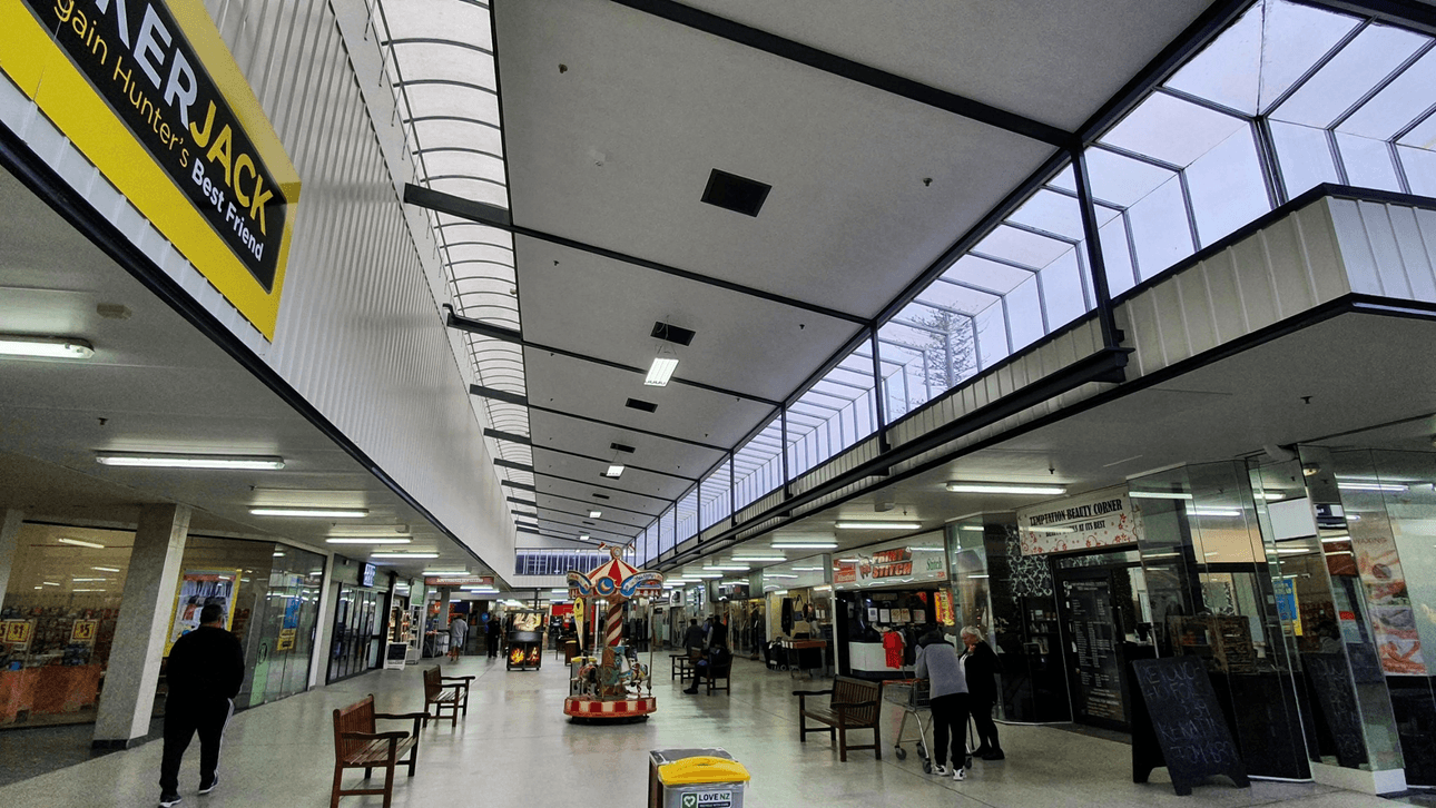 A spacious indoor shopping arcade with a high, transparent ceiling. People are walking and interacting near various shops lining both sides. Wooden benches and a colorful display are positioned along the center corridor.