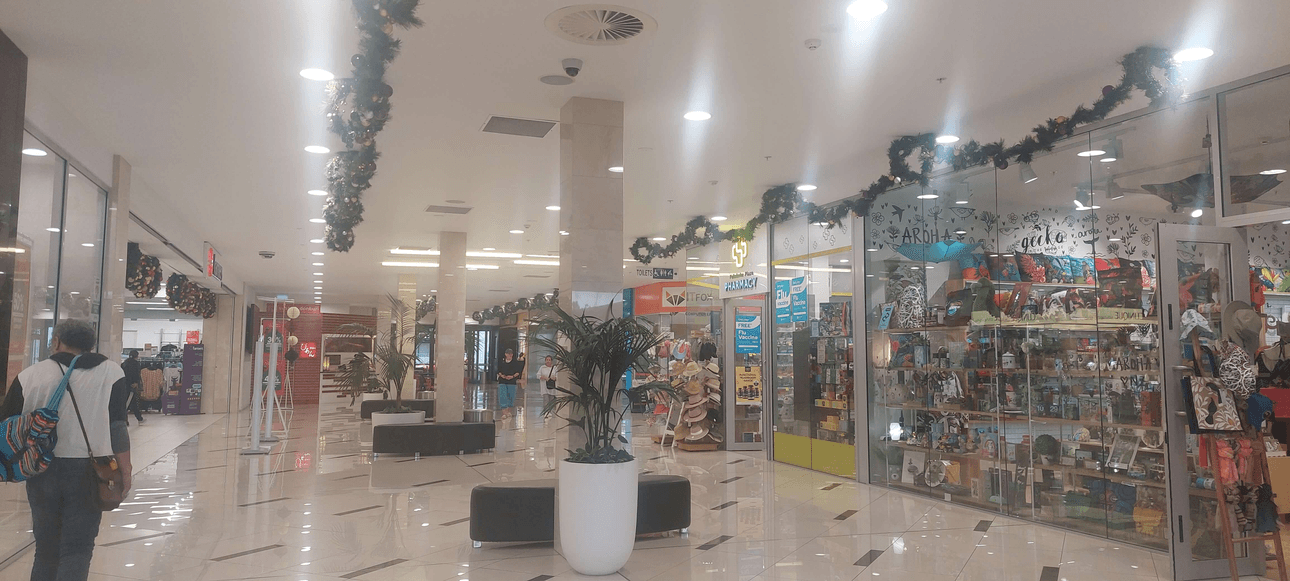 A shopping mall corridor decorated with holiday garlands. Features various stores, including a toy shop with colourful displays and a travel agency. Shoppers walk through the area, and potted plants are placed along the tiled floor.