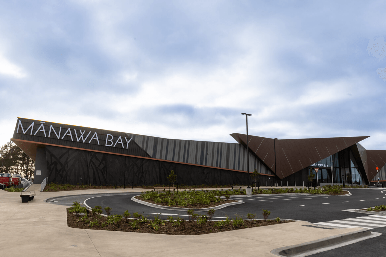 Exterior view of the Mānawa Bay building, featuring modern architecture with angular roofs and large signage. The foreground includes a paved parking area and landscaped greenery under a cloudy sky.