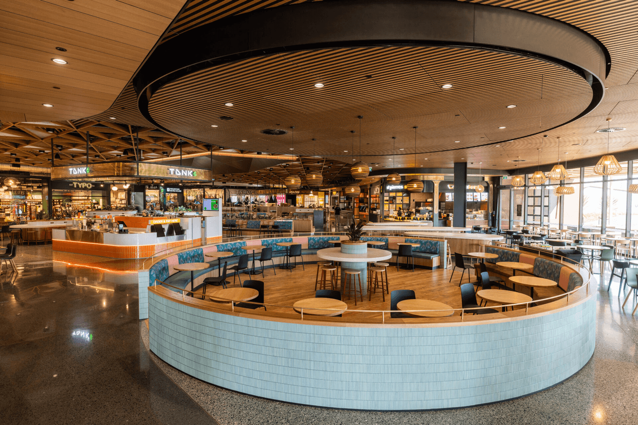 A modern food court featuring various eateries with illuminated signs. Central circular seating area with wooden tables and cushioned benches. Overhead, there are wooden and black circular ceiling designs. The space is spacious with polished floors.