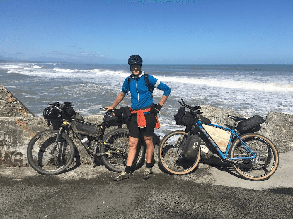 a white man with a blue cycling top on has his face completely shaded by a helmet, standing hammily in front of a loaded bike with a breezy ocean behind him, horsetails getting whipped up behind the waves