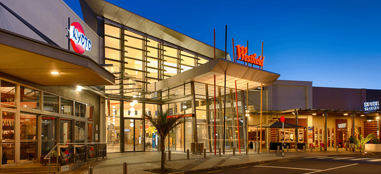 A modern shopping centre entrance at dusk. The building features a large glass facade with the "Westfield" sign illuminated in red. There are outdoor seating areas and palm trees nearby, under a clear evening sky.