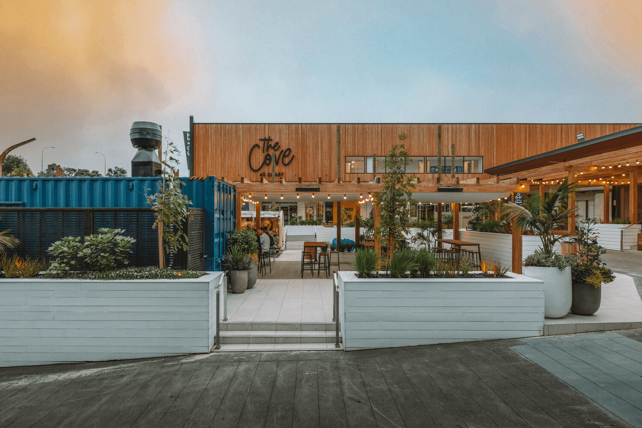 Outdoor dining area with string lights creating a warm ambiance. Wooden pergola and lush greenery surround tables and chairs. People are seated at tables, and a small food trailer is visible in the corner. Cloudy sky overhead.