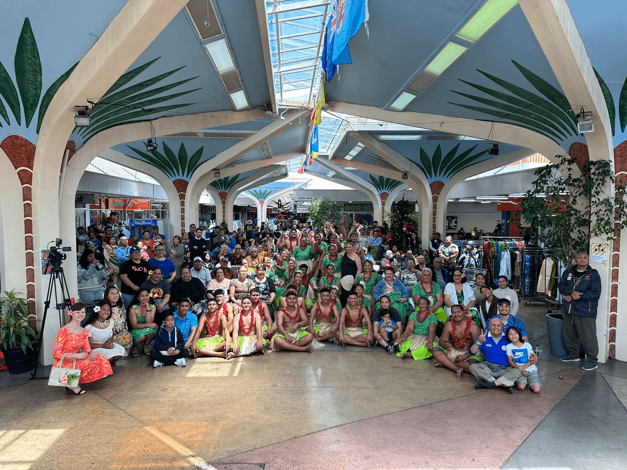 A large group of people, including dancers in traditional Hawaiian attire, pose for a photo in a decorated indoor space with palm tree pillars. The scene is lively and colorful, with various expressions of joy and camaraderie.