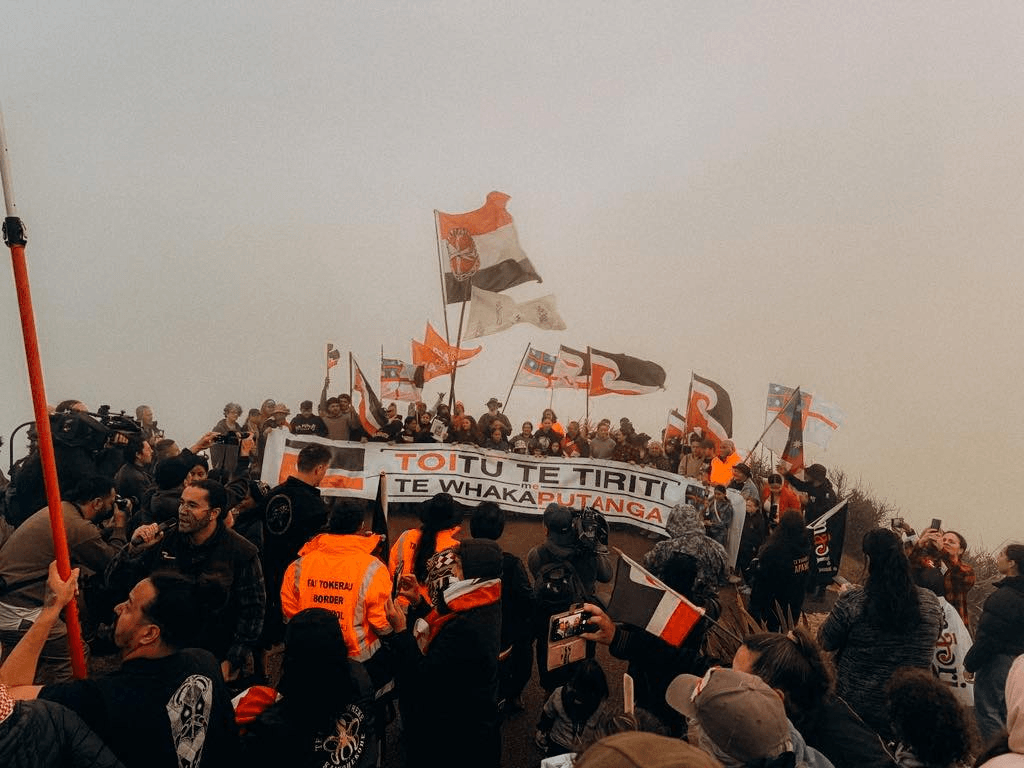 A large group of people are gathered on a hill, holding various flags and a banner that reads "Toitū te Tiriti" and "Te Whaka Putanga." The atmosphere is misty, and several individuals are taking photos or recording the event.