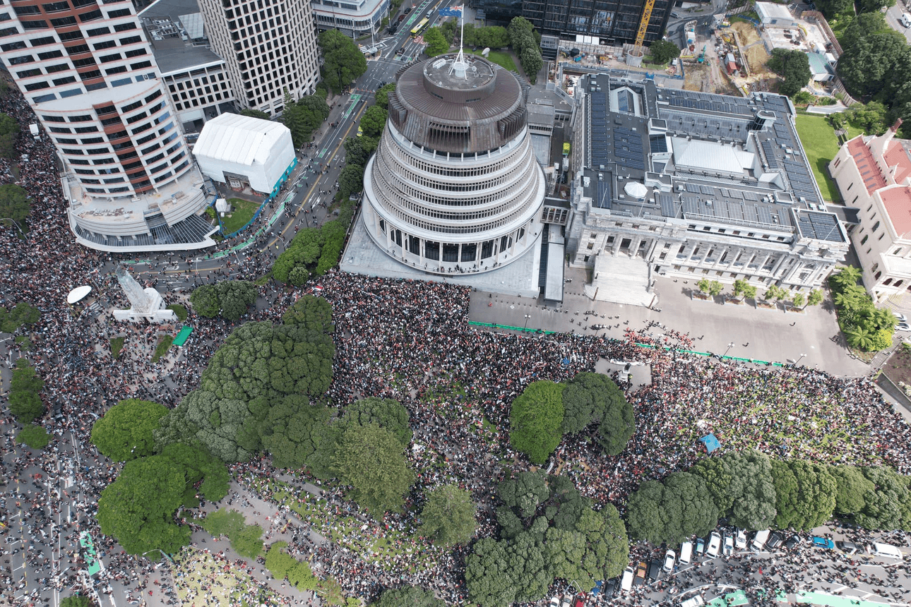 A large crowd gathers around a distinctive circular building with surrounding trees and other structures. The scene is bustling with activity, indicating a significant event or protest. High-rise buildings are visible nearby.