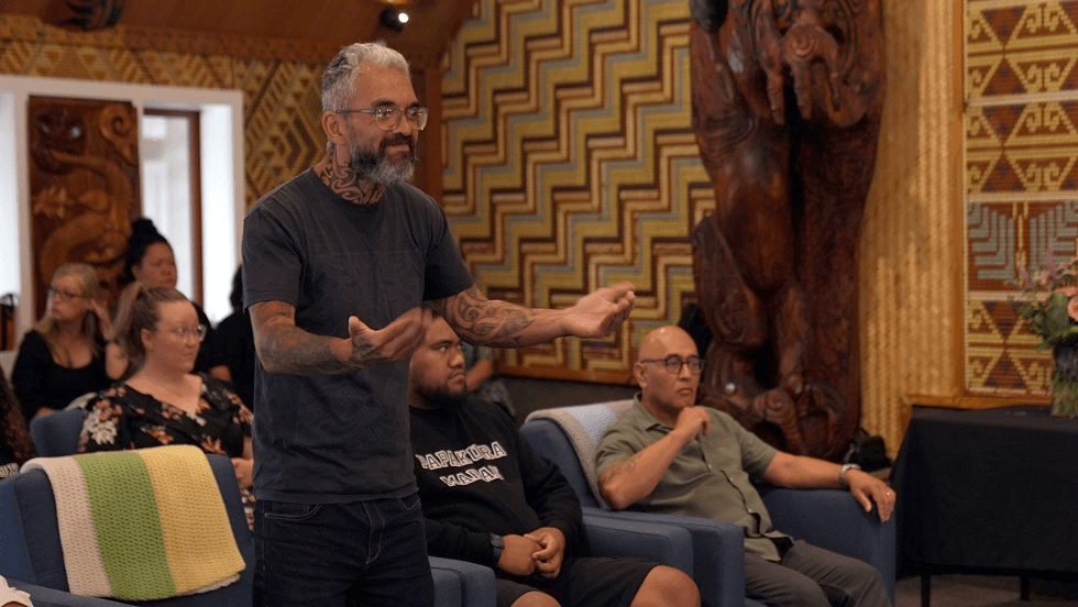 A man with tattoos on his arms and neck is speaking animatedly in a room decorated with traditional carvings and patterns. Several people are seated around him, listening attentively.